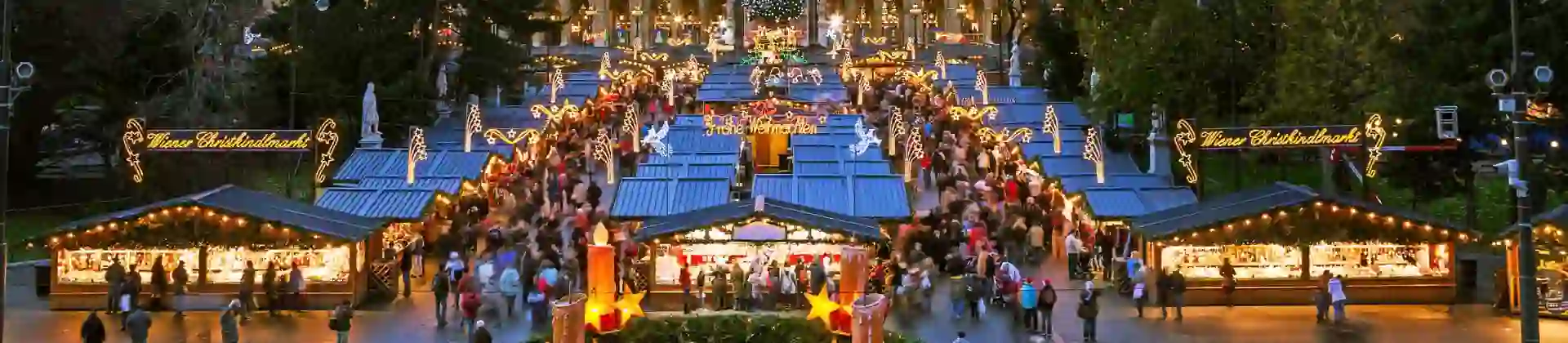 Shot of a gothic city hall which has a tall clock tower in the middle and four smaller towers, two either side, all with spiky turrets. The building has long windows all over and is a gold colour. Below are the Vienna Christmas markets, with four vertical strips of stalls, the centre having a christmas tree at the far end. There are many people shopping, with lights and lit up signs above them.