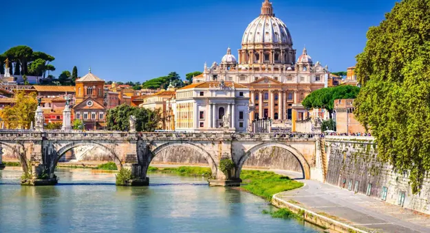 View of St Peter's Basilica in Vatican City, with the River Tiber and the historic Ponte Sant'Angelo bridge in the foreground, on a bright sunny day in Rome, Italy