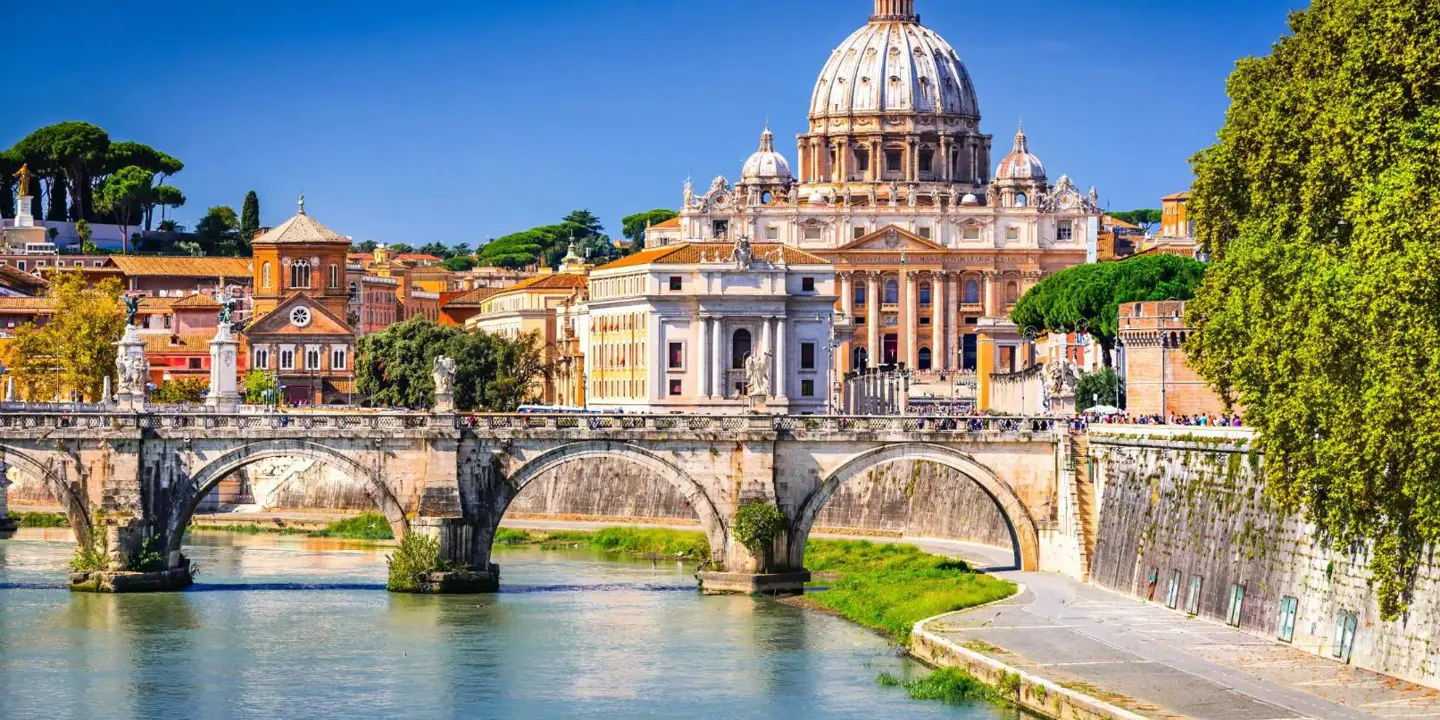 View of St Peter's Basilica in Vatican City, with the River Tiber and the historic Ponte Sant'Angelo bridge in the foreground, on a bright sunny day in Rome, Italy