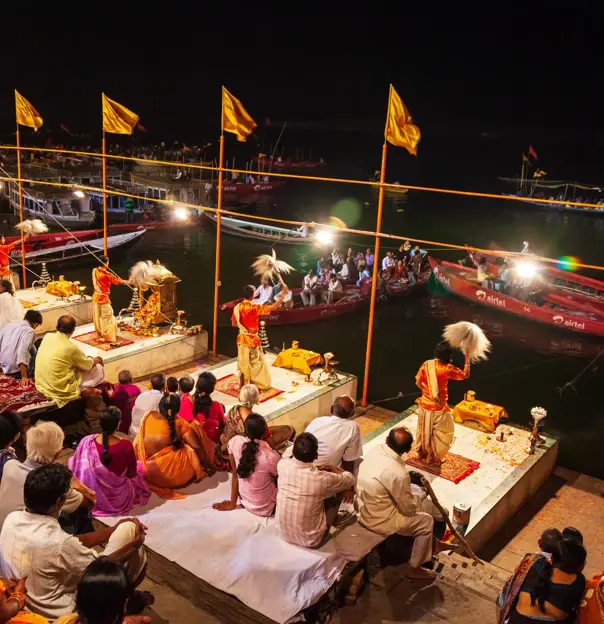 Priests dressed in traditional saffron robes performing the Ganga Aarti ceremony on the ghats of Varanasi at night, surrounded by devotees and illuminated by rows of lights along the riverbank