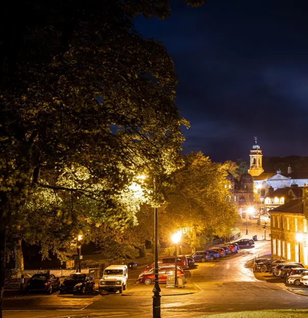 Shot of a road in the town of Buxton at night time, showing trees, houses, parked cars and St John's Church