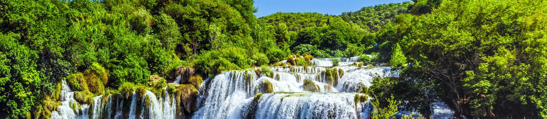 View of a waterfall in Krka National Park, with bright green trees behind it and a blue, clear sky above. Green and blue waters in the forefront.