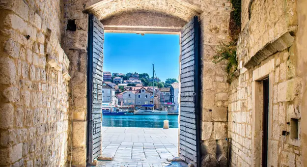 View of a boat on some water, with buildings behind it, through open doors in a stone walkway. A bright blue sky.