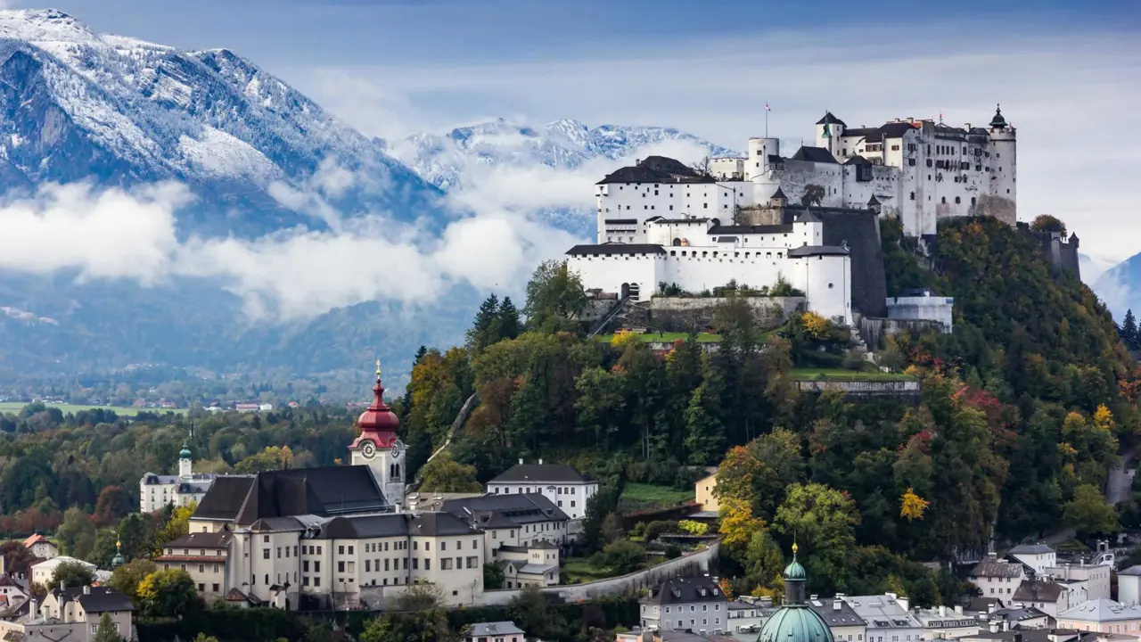 View of the Hohensalzburg Fortress, a white castle with black roofs on top of a grassy hill. At the bottom of the hill is a white large building with a red turret. Behind these is a large mountain with snow on and clouds in front of it.