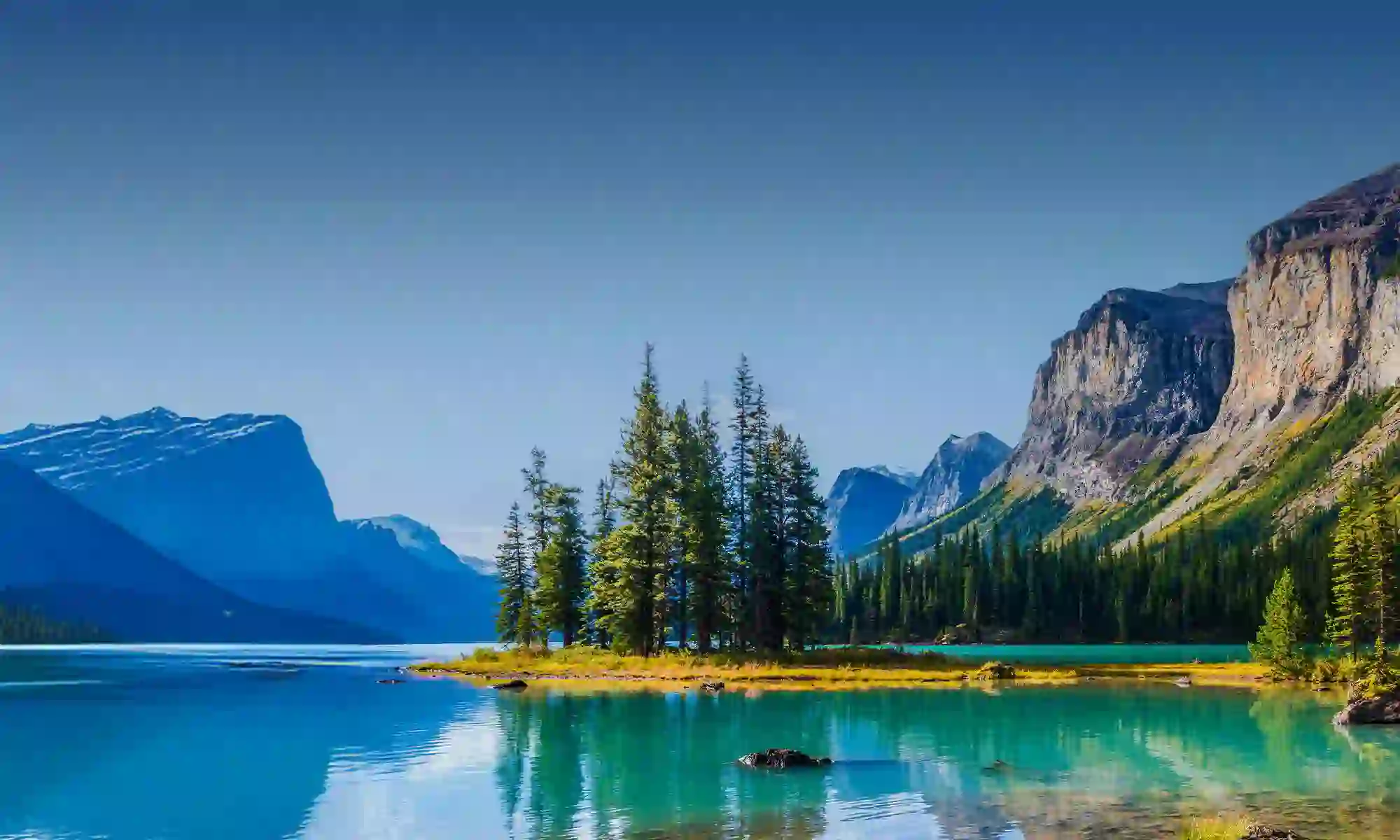 Scenic view of a turquoise lake with a tree-covered island, surrounded by mountains and cliffs under a clear blue sky in Jasper National Park, Canada