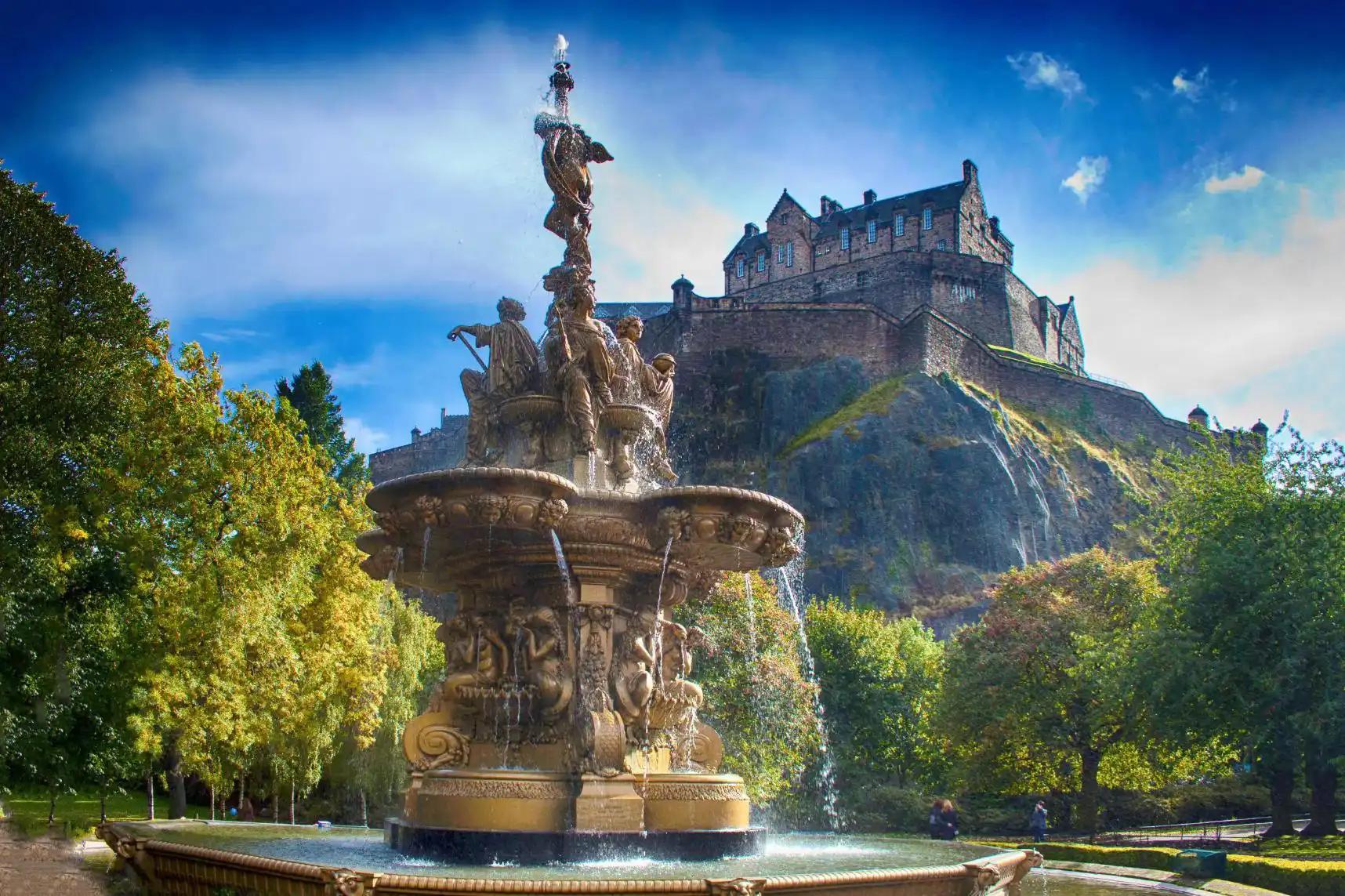 A view of Edinburgh Castle perched high on a rocky hill in the background, with a decorative fountain in the foreground below, surrounded by greenery