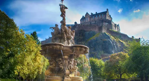 A view of Edinburgh Castle perched high on a rocky hill in the background, with a decorative fountain in the foreground below, surrounded by greenery