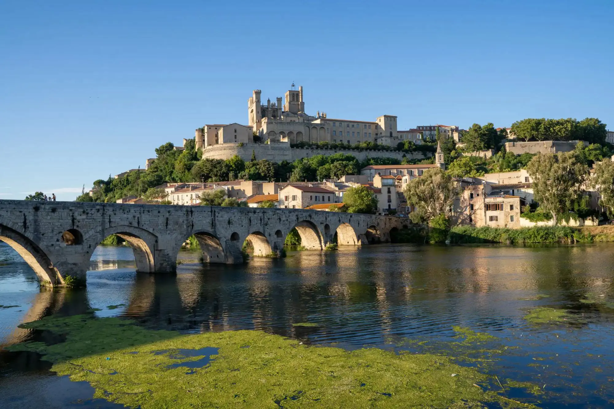 A historic stone bridge crossing a river, leading to the ancient town of Beziers in southern France, with old buildings and a large cathedral on a hill under a clear blue sky