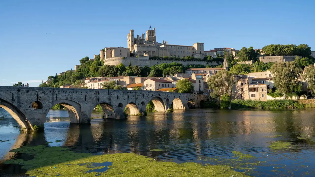 A historic stone bridge crossing a river, leading to the ancient town of Narbonne in southern France, with old buildings and a large cathedral on a hill under a clear blue sky
