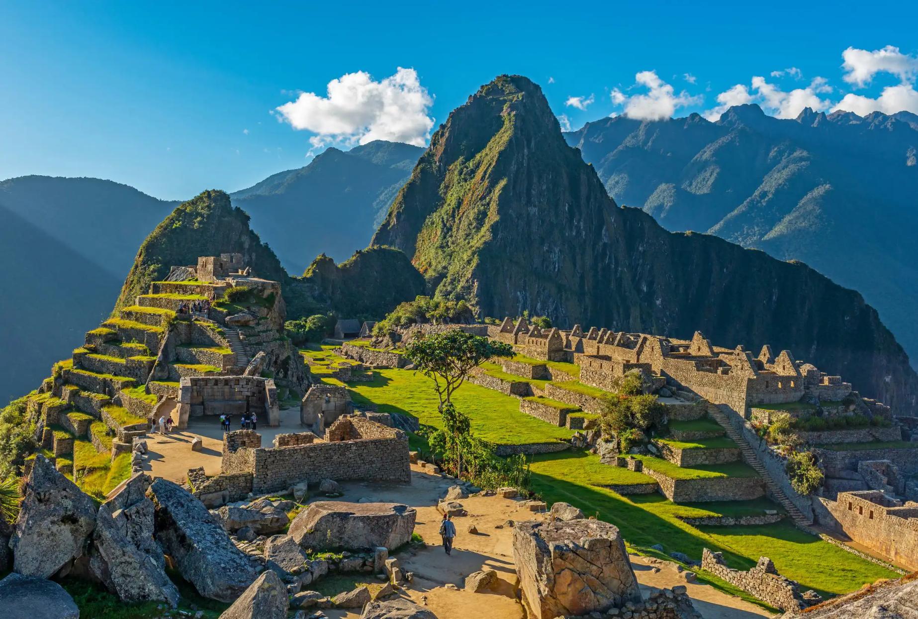 A panoramic view of Machu Picchu in Peru, showing ancient Incan ruins built into a green mountain ridge under a clear blue sky, with Huayna Picchu towering in the background