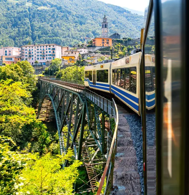 Centovalli Railway bridge in Switzerland with a train crossing, surrounded by mountainous countryside, and a town visible in the distance