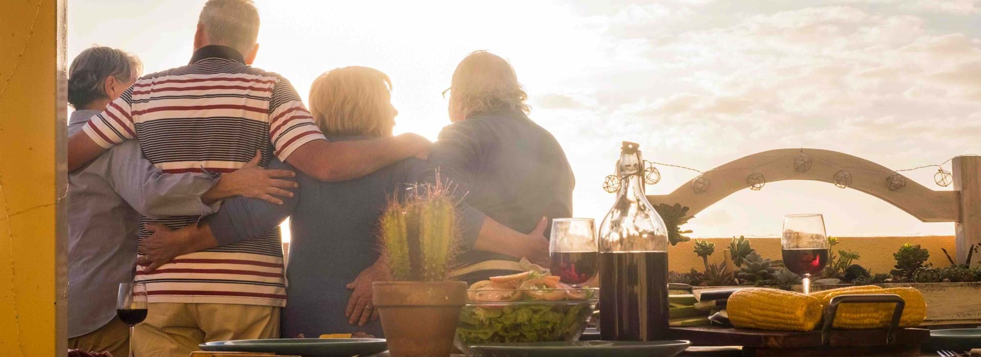 4 friends looking out over thier balcony with a table that has wine & food on it behind them 