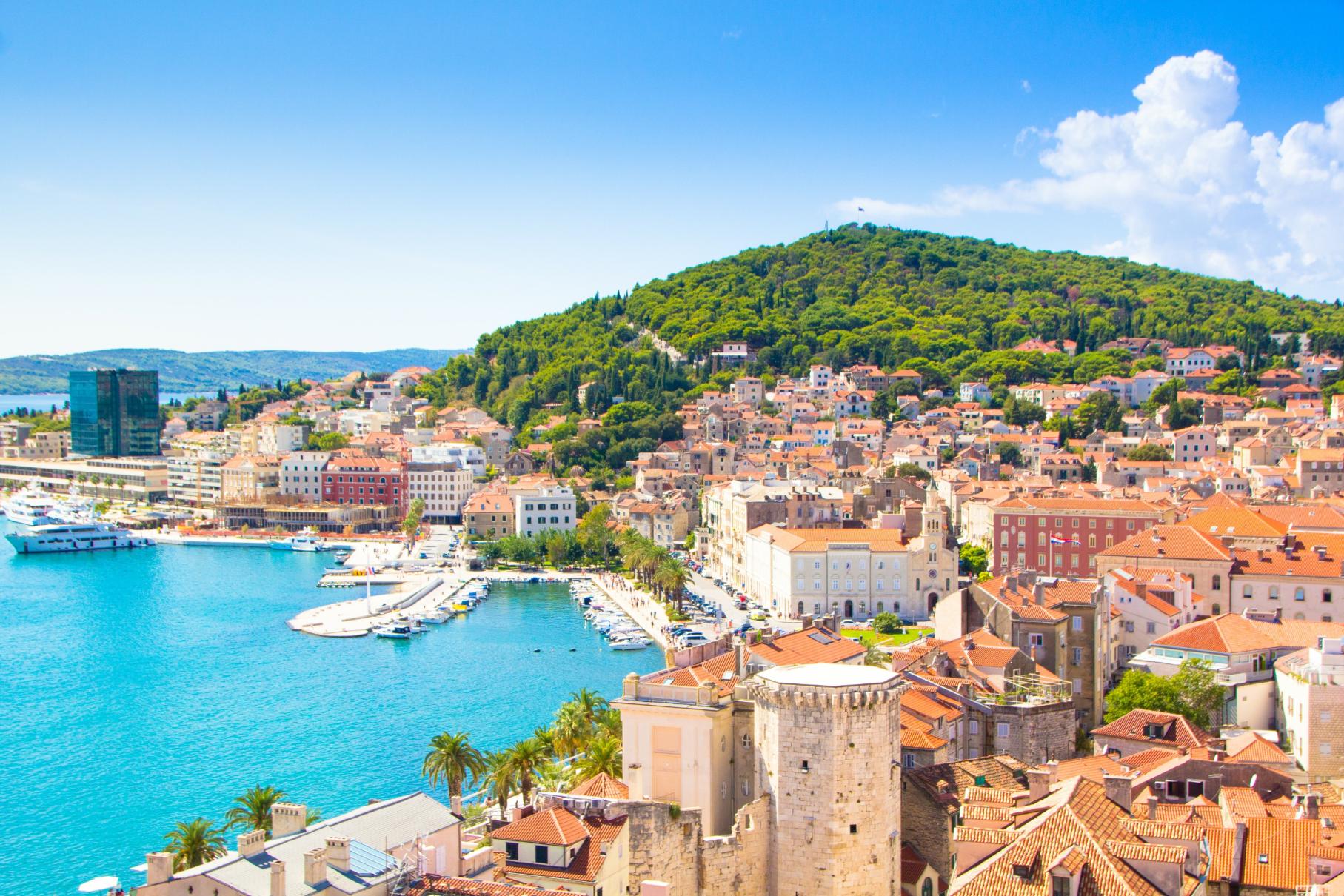A panoramic view of Split, Croatia, showing the turquoise Adriatic Sea, a bustling harbour with yachts, and the terracotta rooftops of Old Town at the foot of lush green Marjan Hill