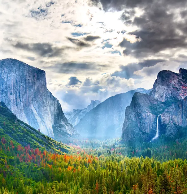 Yosemite National Park in California with towering granite cliffs, green forested areas, and cloudy skies with sun rays breaking through