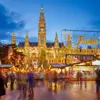 Shot of Vienna's gothic city hall which has one tall clock tower and four smaller towers, two on each side, all with pointy turrets. The building is a gold colour and is in front of a bright blue night sky. In front, is a Christmas Market, showing strips of cabin-like stalls and a variety of lit up Christmas decorations, one in the forefront reading 'Merry Christmas' in Austrian. People can be seen, although blurry as they are moving around. 