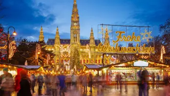 Shot of Vienna's gothic city hall which has one tall clock tower and four smaller towers, two on each side, all with pointy turrets. The building is a gold colour and is in front of a bright blue night sky. In front, is a Christmas Market, showing strips of cabin-like stalls and a variety of lit up Christmas decorations, one in the forefront reading 'Merry Christmas' in Austrian. People can be seen, although blurry as they are moving around. 