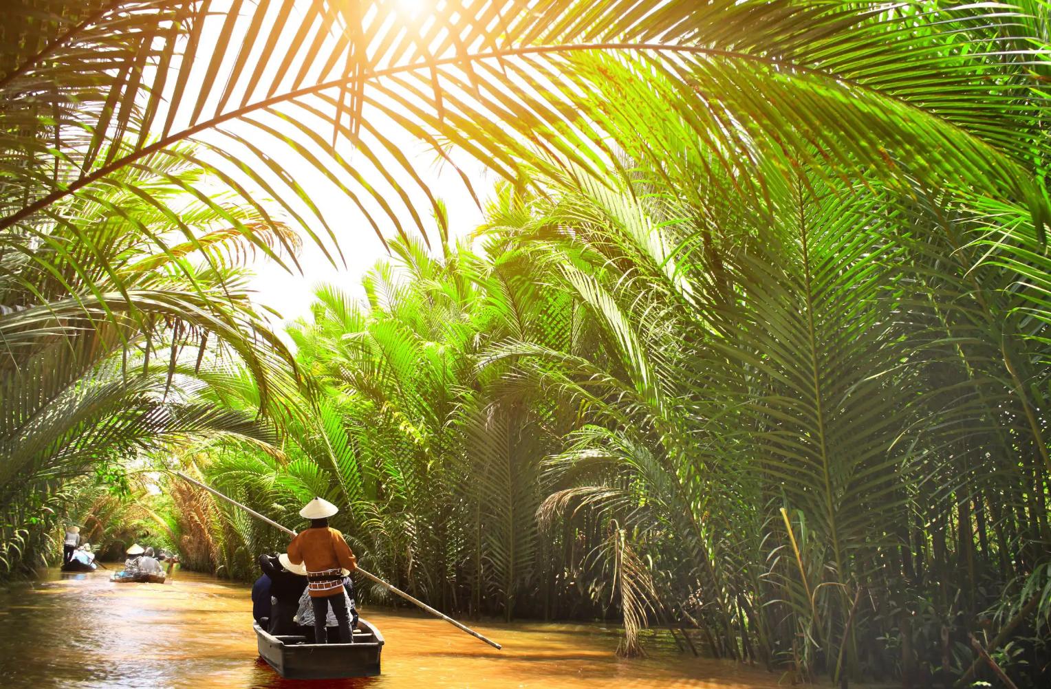 Small wooden boats glide through a palm-lined canal off the Mekong River in Thailand, with a person in a conical hat steering under filtered sunlight