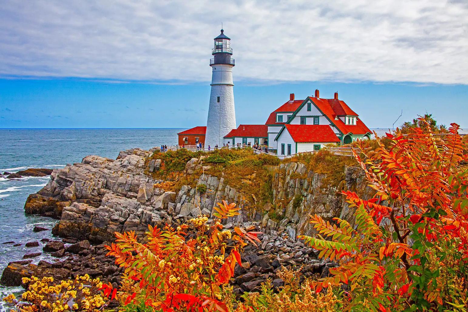White lighthouse and red-roofed buildings on a rocky coast in Portland, with autumn foliage in the foreground