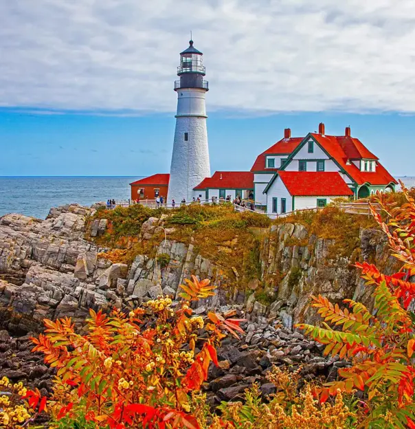 White lighthouse and red-roofed buildings on a rocky coast in Portland, with autumn foliage in the foreground