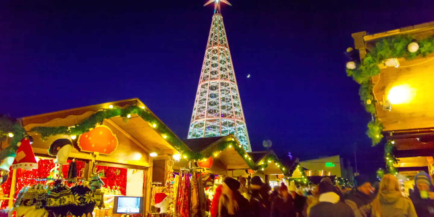 View of a Christmas market, with wooden, cabin-like stalls in the forefront, selling winter hats, dream catchers and scarves. The roofs are lined with green leaf-like garlands and fairy lights. Behind these is a Christmas tree metal structure, with a large, lit up star on top, in front of a navy blue night sky.