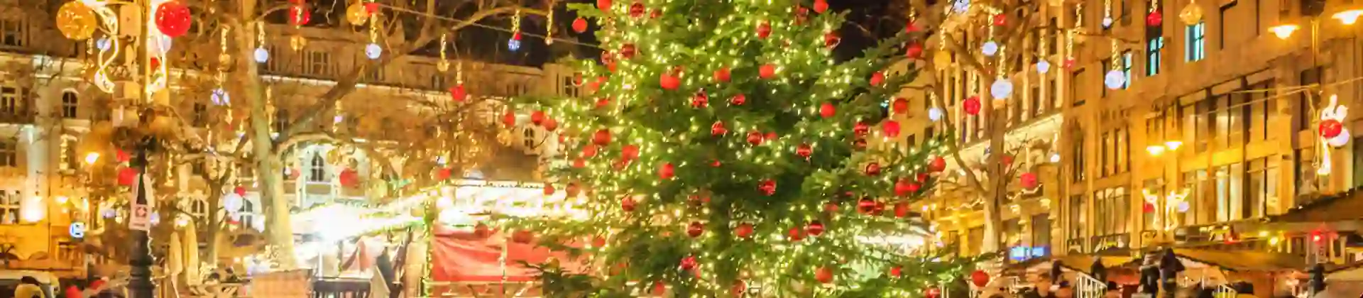 A square in Budapest at Christmas, in the front, a 'Budapest' sign, with a Christmas tree with lights and red baubles behind. Behind this, lit up decorations with hanging baubles, strung across the square.