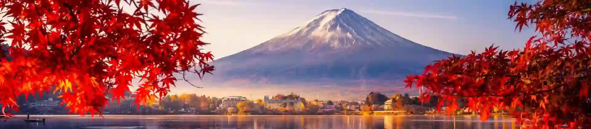 Mount Fuji reflected in Lake Kawaguchi, framed by vivid red autumn leaves, with a sunlit lakeside town between the water and the mountain