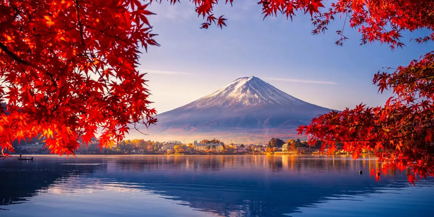 Mount Fuji reflected in Lake Kawaguchi, framed by vivid red autumn leaves, with a sunlit lakeside town between the water and the mountain