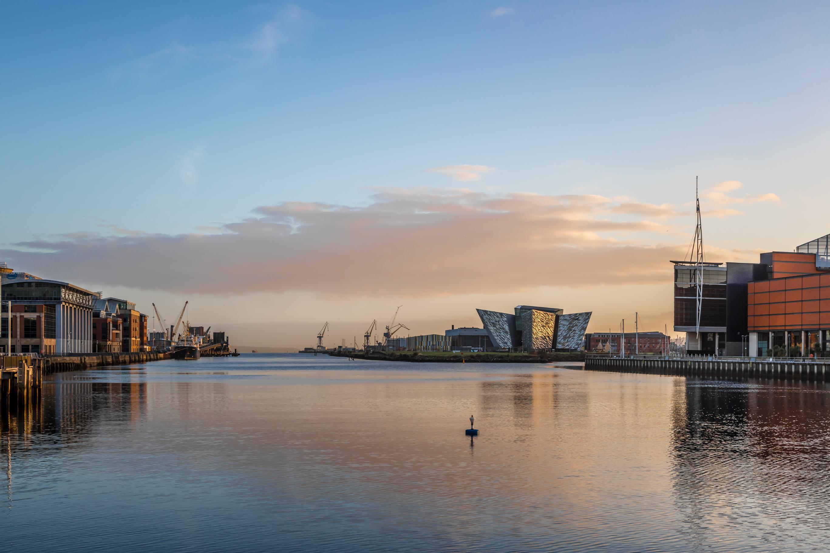 Titanic Belfast museum, shaped like ship hulls, sits on the waterfront surrounded by old shipyard cranes and calm water at sunset