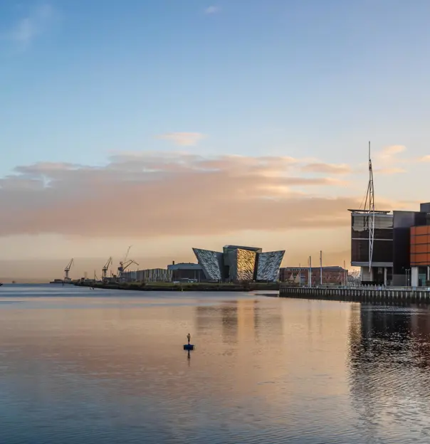Titanic Belfast museum, shaped like ship hulls, sits on the waterfront surrounded by old shipyard cranes and calm water at sunset
