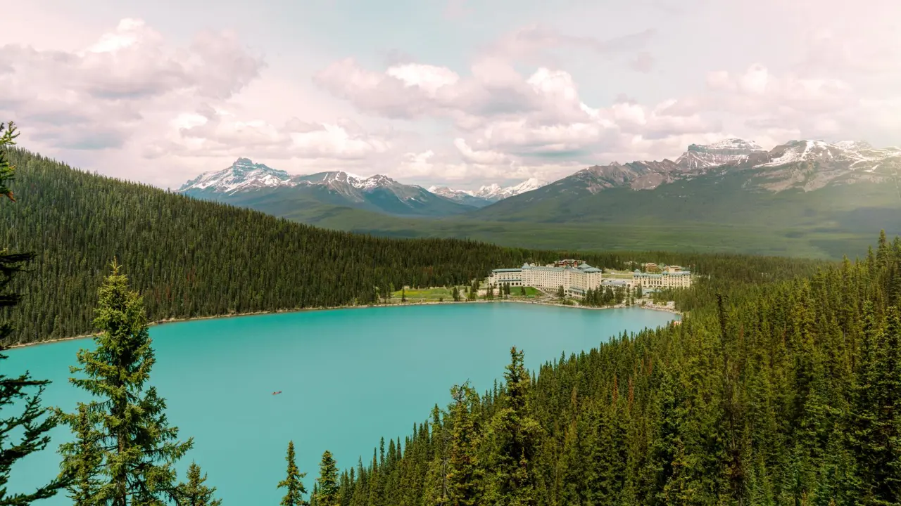 Fairmont Chateau, Lake Louise next to a turquoise lake which is surrounded by dense pine forest, under a bright sky