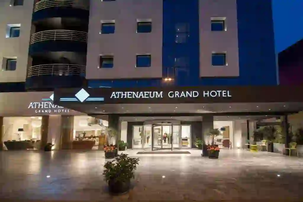 Front entrance of the Athenaeum Grand Hotel at night, with modern signage, glass doors, and potted plants