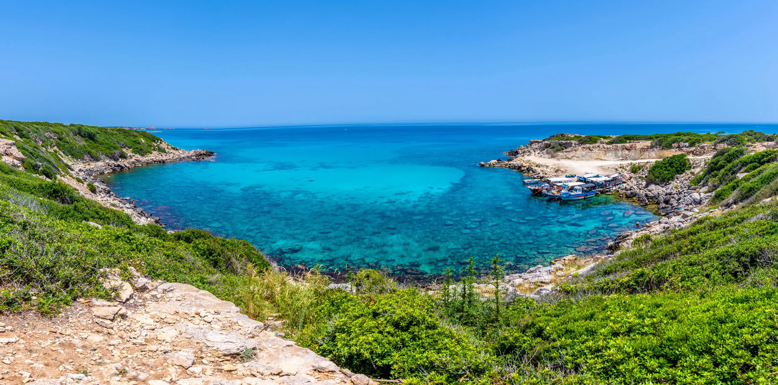 View of the sea from land, the edge being a circular shape with some boats docked to the right. The land has some grass and the sky is blue.