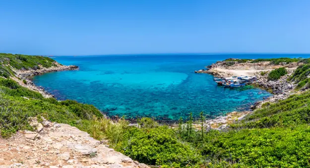 View of the sea from land, the edge being a circular shape with some boats docked to the right. The land has some grass and the sky is blue.