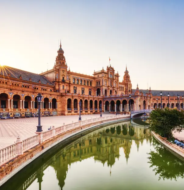 Wide view of Plaza de España in Seville, featuring its grand semicircular building, ornate tilework, and the small river with a bridge crossing it in the distance