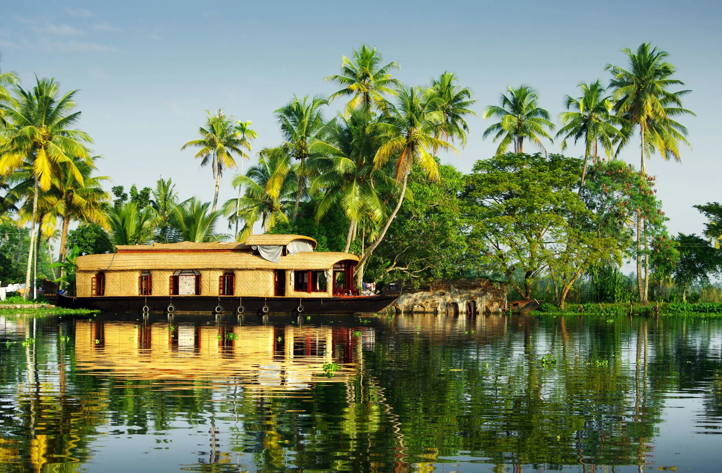 A light brown wicker houseboat on calm water in Kerala, with palm trees on the banks and clear blue skies overhead