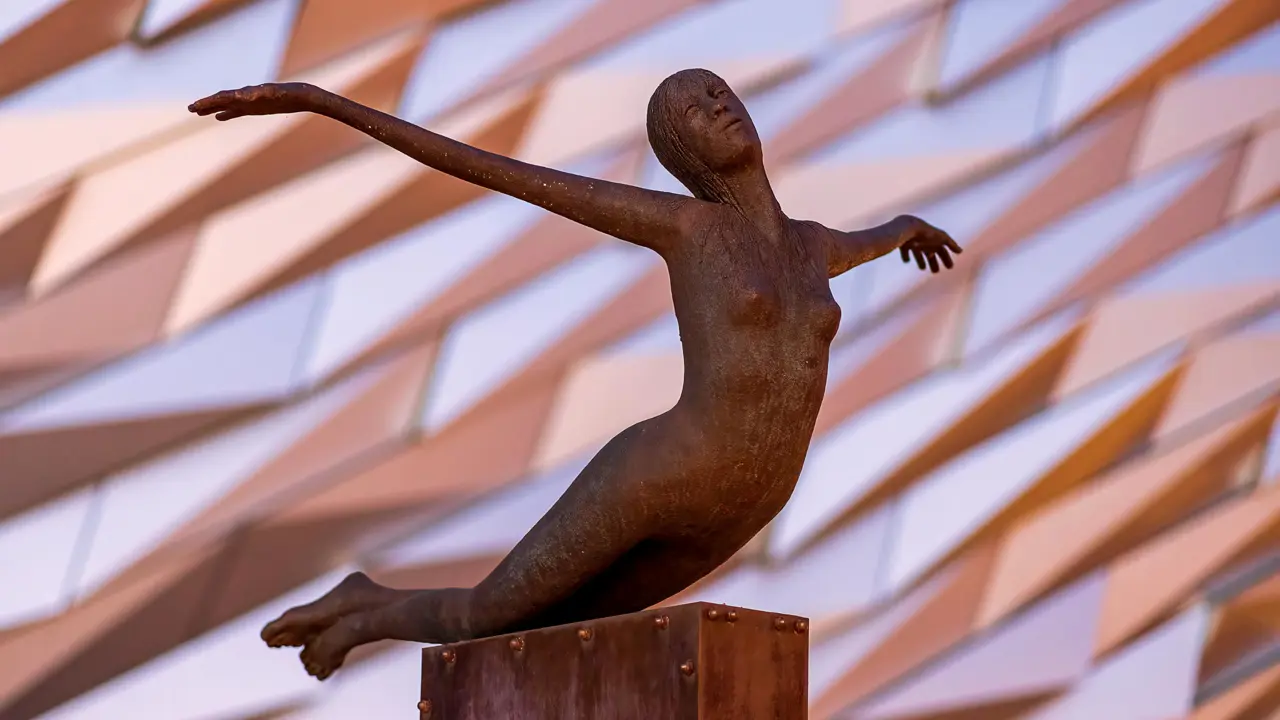 Bronze sculpture of a woman with arms outstretched in a diving pose, set against the angular facade of the Titanic Belfast museum