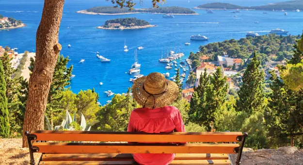 Back of a woman wearing a sun hat, sat on a bench looking out to sea. Variety of boats on the water, and forested land to the right.