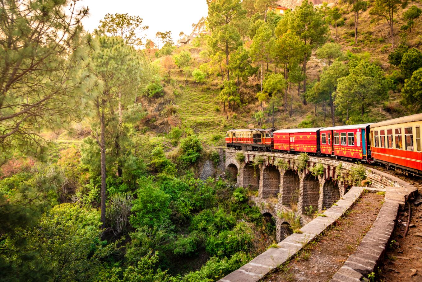 The heritage toy train crossing a railway bridge on the Kalka–Shimla route, surrounded by lush green hills and trees