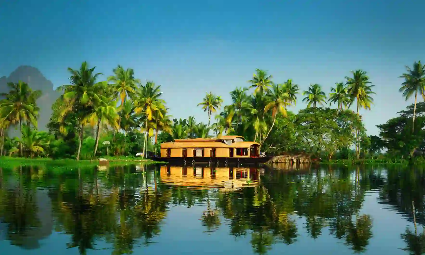 A traditional wooden houseboat floats on calm waters surrounded by lush green palm trees under a clear blue sky in Kerala, India