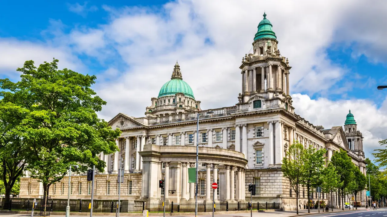 Bright daytime view of Belfast City Hall with its grand columns, domes, and green copper roof, surrounded by trees and traffic lights