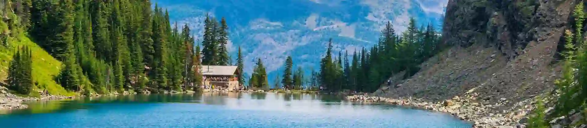 A clear turquoise lake with gentle ripples in Banff National Park, Alberta, Canada, bordered by lush green pine forests and rolling hills, with a small cabin situated at the far end of the lake