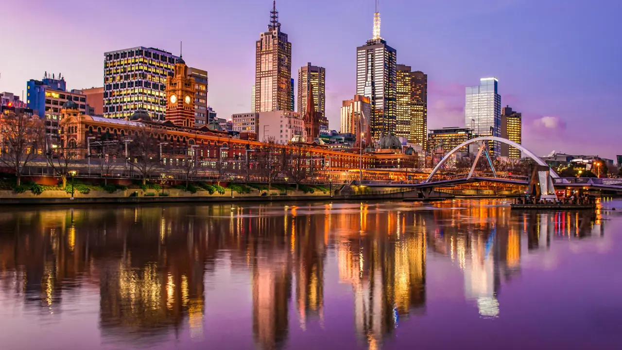 A panoramic view of Melbourne, Victoria at dusk: the shimmering waters of the Yarra River in the foreground, iconic city-skyscrapers rising in the background, and soft pastel hues in the evening sky.