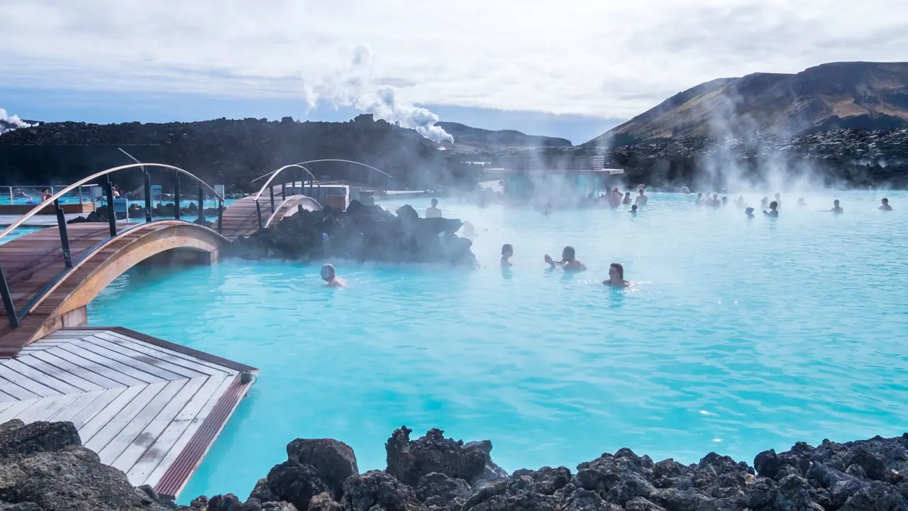 Geothermal lagoon in Iceland - bright blue water with steam coming off, surrounded by black rock. People swimming.