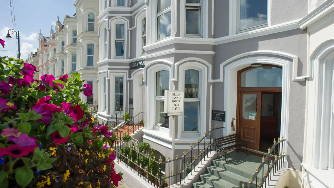Front entrance of the Ellan Vannin Hotel, a traditional building with bay windows, grey and white exterior, decorative railings, and vibrant purple flowers in the foreground
