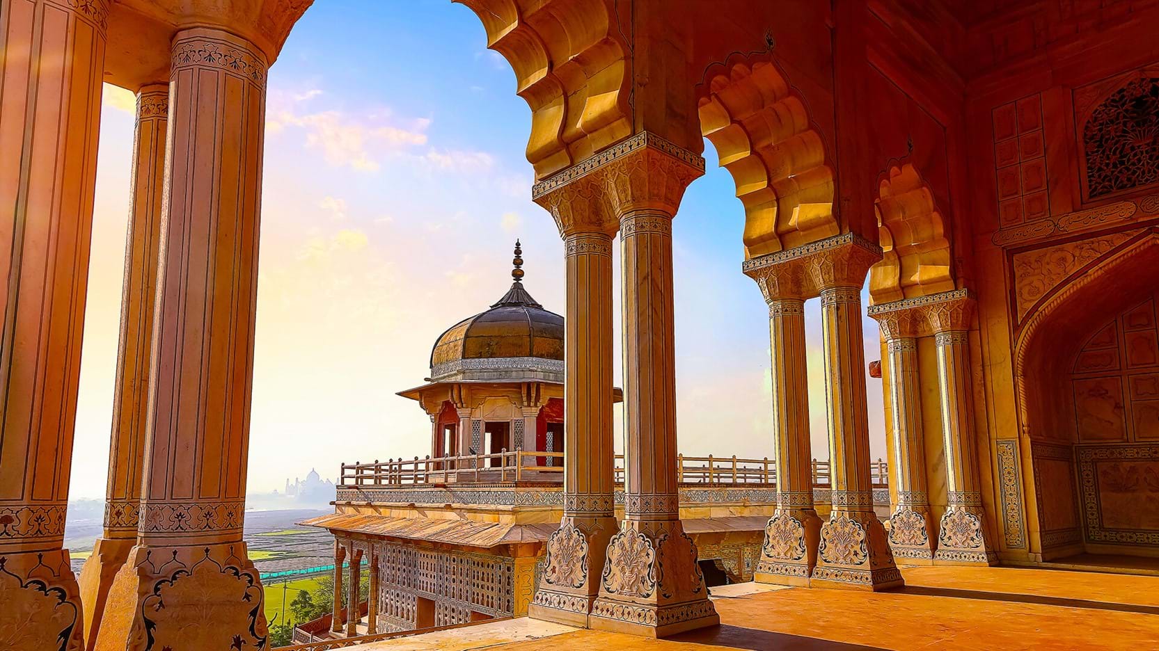 A view through ornate sandstone arches at Agra Fort, overlooking a domed pavilion and distant landscape in warm evening light