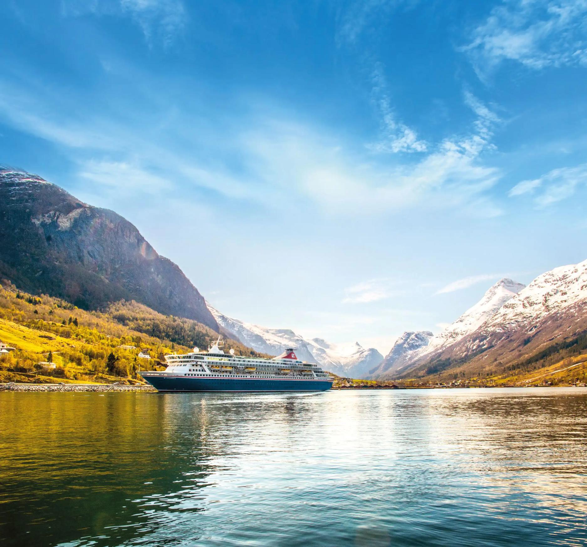 The Balmoral cruise ship sails through a tranquil fjord in Norway, surrounded by steep, snow-capped mountains and lush, forested hills beneath a bright blue sky
