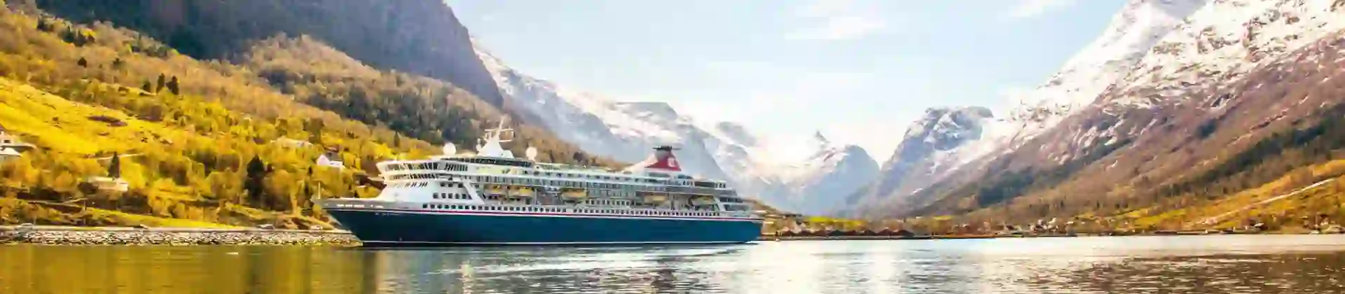 The Balmoral cruise ship sails through a tranquil fjord in Norway, surrounded by steep, snow-capped mountains and lush, forested hills beneath a bright blue sky