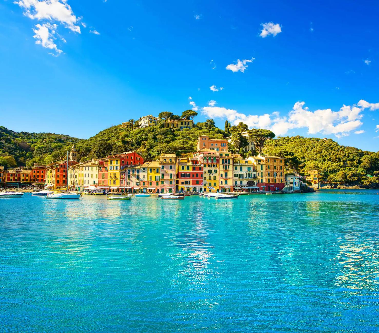 Picturesque view of Portofino, Italy, featuring colourful harbourfront buildings, boats gently floating in the clear blue water, and lush green hills in the background