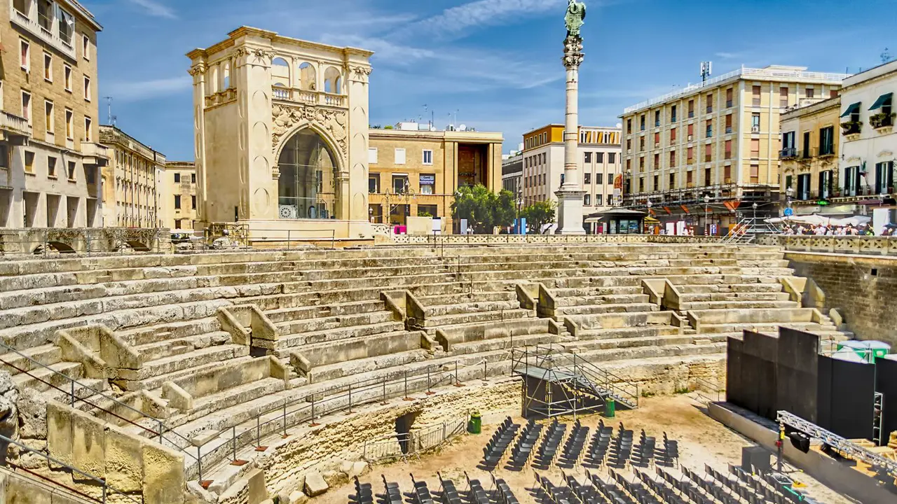 A Roman amphitheatre with stone seating in Sant'Oronzo Square, Lecce, under a clear blue sky, surrounded by modern buildings and event chairs on the ground