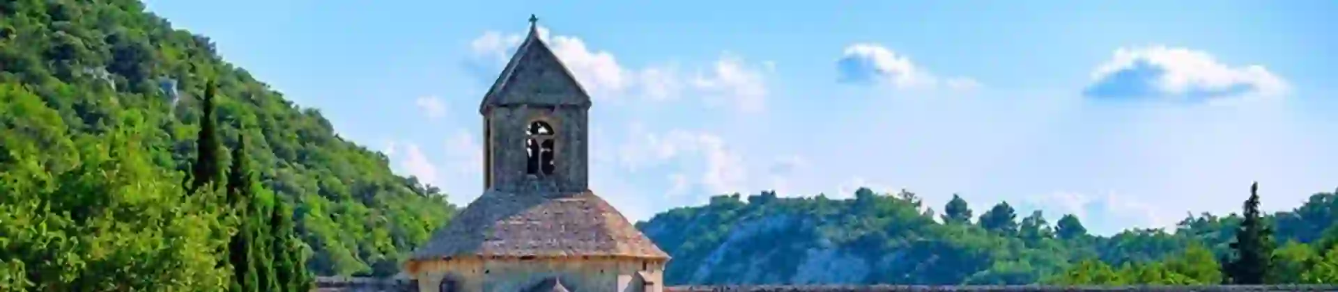 View of a wide, grey, stone church with rows of lavender in front of it, all behind a grey bricked wall. To the left, a grassy hill and some trees